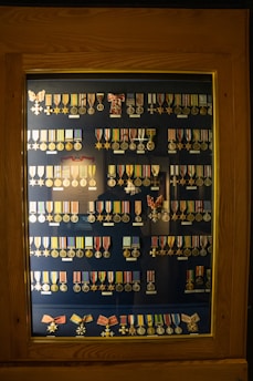 A wooden display case contains rows of various military medals and ribbons organized neatly. The medals are of different designs and colors, primarily gold and silver, and some feature stars or crosses. Many have colored ribbons attached, displaying shades of red, blue, yellow, and green. The background is a dark blue, enhancing the medals' visibility.