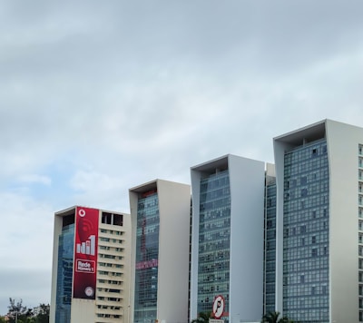 Several modern office buildings with large glass windows and white facades are set against a cloudy sky. The leftmost building displays a large red vertical billboard promoting a network service. Palm trees are visible at the base of the structures, adding a touch of greenery.