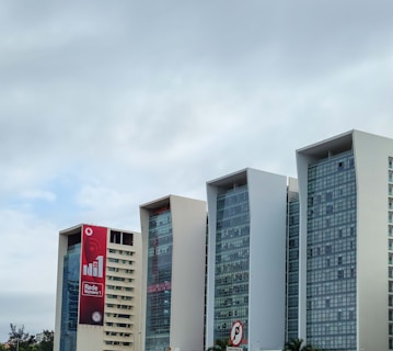 Several modern office buildings with large glass windows and white facades are set against a cloudy sky. The leftmost building displays a large red vertical billboard promoting a network service. Palm trees are visible at the base of the structures, adding a touch of greenery.