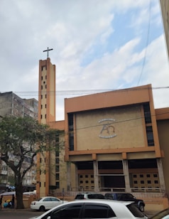 A large church building with a tall tower featuring a cross at the top. The facade is a mix of beige and brown tones, with an emblem and some text above the entrance. Several cars are parked in front of the building, and there is a tree on the left side. The sky is partly cloudy with patches of blue visible.