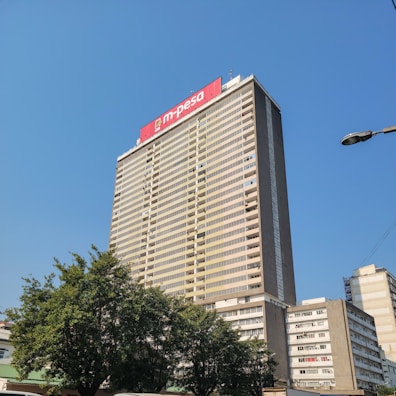 A tall office building rises against a clear blue sky. The facade is rectangular with numerous windows, and a prominent red sign with 'm-pesa' is displayed at the top. Lush green trees are visible at the base of the building, adding a touch of nature to the urban setting. Nearby, other smaller buildings are also visible.