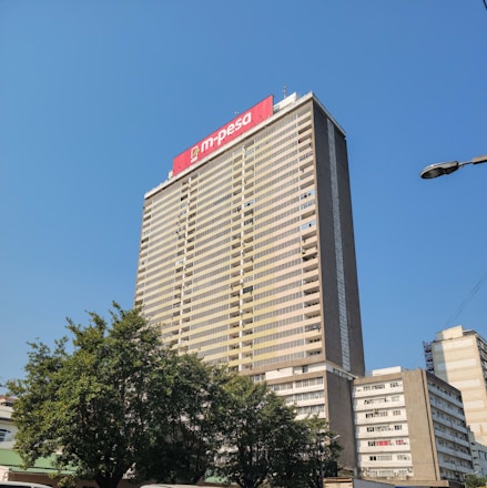 A tall office building rises against a clear blue sky. The facade is rectangular with numerous windows, and a prominent red sign with 'm-pesa' is displayed at the top. Lush green trees are visible at the base of the building, adding a touch of nature to the urban setting. Nearby, other smaller buildings are also visible.