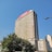 A tall office building rises against a clear blue sky. The facade is rectangular with numerous windows, and a prominent red sign with 'm-pesa' is displayed at the top. Lush green trees are visible at the base of the building, adding a touch of nature to the urban setting. Nearby, other smaller buildings are also visible.