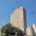 A tall office building rises against a clear blue sky. The facade is rectangular with numerous windows, and a prominent red sign with 'm-pesa' is displayed at the top. Lush green trees are visible at the base of the building, adding a touch of nature to the urban setting. Nearby, other smaller buildings are also visible.