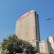 A tall office building rises against a clear blue sky. The facade is rectangular with numerous windows, and a prominent red sign with 'm-pesa' is displayed at the top. Lush green trees are visible at the base of the building, adding a touch of nature to the urban setting. Nearby, other smaller buildings are also visible.