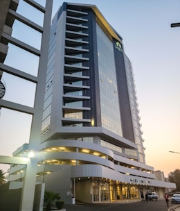 A tall and modern office building with a glass facade, featuring multiple balconies and a distinctive angled roof. It is situated in an urban area with some cars parked outside and a few people walking nearby. The structure appears sleek and well-maintained, with a sign reading 'MAPUTO BUSINESS TOWER' at the entrance.