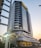 A tall and modern office building with a glass facade, featuring multiple balconies and a distinctive angled roof. It is situated in an urban area with some cars parked outside and a few people walking nearby. The structure appears sleek and well-maintained, with a sign reading 'MAPUTO BUSINESS TOWER' at the entrance.