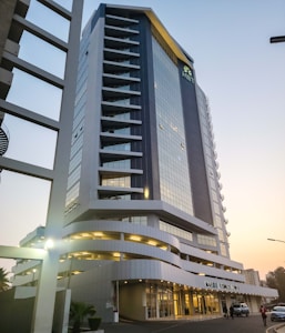 A tall and modern office building with a glass facade, featuring multiple balconies and a distinctive angled roof. It is situated in an urban area with some cars parked outside and a few people walking nearby. The structure appears sleek and well-maintained, with a sign reading 'MAPUTO BUSINESS TOWER' at the entrance.