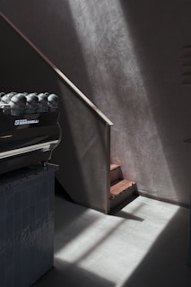 Warm sunlight filters through large windows onto a sleek espresso machine surrounded by dark wood and matte black accents.