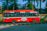 A red bus is traveling quickly along a road with a blur effect indicating speed. It features zero emission branding on the side. Inside the bus, passengers are visible through the windows. The background shows greenery with trees and a railing, suggesting an urban or suburban setting.