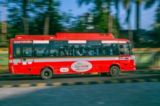 A red bus is traveling quickly along a road with a blur effect indicating speed. It features zero emission branding on the side. Inside the bus, passengers are visible through the windows. The background shows greenery with trees and a railing, suggesting an urban or suburban setting.