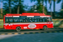A red bus is traveling quickly along a road with a blur effect indicating speed. It features zero emission branding on the side. Inside the bus, passengers are visible through the windows. The background shows greenery with trees and a railing, suggesting an urban or suburban setting.