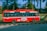 A red bus is traveling quickly along a road with a blur effect indicating speed. It features zero emission branding on the side. Inside the bus, passengers are visible through the windows. The background shows greenery with trees and a railing, suggesting an urban or suburban setting.