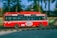 A red bus is traveling quickly along a road with a blur effect indicating speed. It features zero emission branding on the side. Inside the bus, passengers are visible through the windows. The background shows greenery with trees and a railing, suggesting an urban or suburban setting.