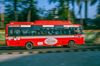 A red bus is traveling quickly along a road with a blur effect indicating speed. It features zero emission branding on the side. Inside the bus, passengers are visible through the windows. The background shows greenery with trees and a railing, suggesting an urban or suburban setting.