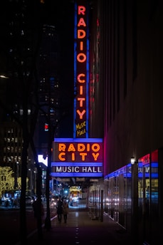 Neon signs in bright red and blue lights illuminate the words 'Radio City Music Hall' on a building's facade at night. The scene shows a bustling urban street with silhouettes of people walking and vehicles, including brightly lit cars, moving along the road.
