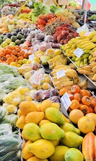 A vibrant market stall displaying a diverse assortment of fresh fruits and vegetables. The produce includes bananas, papayas, avocados, persimmons, and exotic rambutan, neatly arranged in bundles and bags. Signs with handwritten labels indicate the names and prices of various items. The variety of fruits showcases a mixture of colors and textures, enhancing the appeal of this lively market setting.