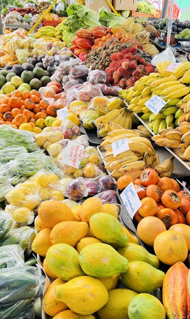 A vibrant market stall displaying a diverse assortment of fresh fruits and vegetables. The produce includes bananas, papayas, avocados, persimmons, and exotic rambutan, neatly arranged in bundles and bags. Signs with handwritten labels indicate the names and prices of various items. The variety of fruits showcases a mixture of colors and textures, enhancing the appeal of this lively market setting.