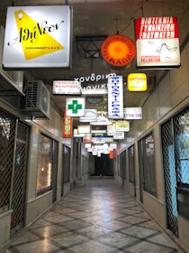 A narrow, indoor passageway is lined with various illuminated signs in different colors and languages, hanging from the ceiling. The corridor is flanked by closed, gated shops with a tiled floor.