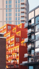 A vibrant urban scene with a mix of architectural styles, featuring a prominent orange-red building that stands out against taller, more muted modern skyscrapers. The orange building has balcony railings and many windows, suggesting residential use, while the adjacent buildings have glass facades.