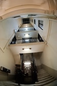 An interior view of a multi-story building with a classic, vintage elevator at the center. The elevator is enclosed in a metal grille and is surrounded by several floors with black railings. Bright lights illuminate the area, highlighting the architectural details of the staircases that wind around the elevator shaft. A person is visible standing near the elevator.