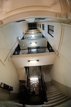 An interior view of a multi-story building with a classic, vintage elevator at the center. The elevator is enclosed in a metal grille and is surrounded by several floors with black railings. Bright lights illuminate the area, highlighting the architectural details of the staircases that wind around the elevator shaft. A person is visible standing near the elevator.