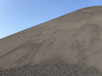 A steep slope covered in dark, fine volcanic ash under a clear blue sky.