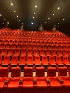 A large, empty movie theater with rows of red recliner seats arranged in a gradient pattern facing the front. Soft ambient lighting casts a warm glow, creating a cozy atmosphere.