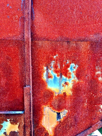 A close-up view of a rusted metal surface with various shades of red and orange corrosion. Patches of blue and yellow are visible where the rust has flaked away, creating an abstract and textured pattern. The surface consists of old panels joined together, highlighting the effects of oxidation.