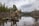 A couple is walking on a log in a serene forest setting, surrounded by a calm lake and tall evergreen trees. Snow-capped mountains are visible in the background under a cloudy sky.