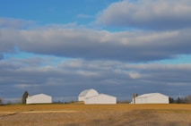 A rural landscape with several white farm buildings, including a barn and a silo, set against a backdrop of a vast, partly cloudy sky. The foreground features a field with dry grass and rolling terrain.