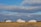A rural landscape with several white farm buildings, including a barn and a silo, set against a backdrop of a vast, partly cloudy sky. The foreground features a field with dry grass and rolling terrain.