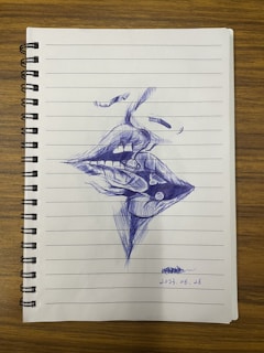 A ballpoint pen drawing on a lined notebook page depicts abstract, surrealistic intertwined lips and facial features. The drawing is detailed with cross-hatching and shading, suggesting depth and texture.