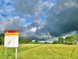 A warning sign stands in a grassy field with power lines stretching across the sky, framed by dense green foliage. Dark storm clouds loom overhead, contrasting with patches of blue sky.