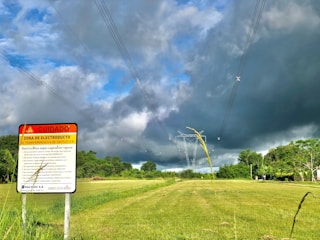 A warning sign stands in a grassy field with power lines stretching across the sky, framed by dense green foliage. Dark storm clouds loom overhead, contrasting with patches of blue sky.