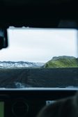 A rugged, volcanic landscape is viewed from inside a vehicle, with a dark, dirt road leading towards distant mountains. Snow-capped peaks contrast with lush, green hills under a pale, overcast sky.