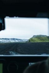 A rugged, volcanic landscape is viewed from inside a vehicle, with a dark, dirt road leading towards distant mountains. Snow-capped peaks contrast with lush, green hills under a pale, overcast sky.