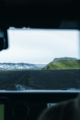 A rugged, volcanic landscape is viewed from inside a vehicle, with a dark, dirt road leading towards distant mountains. Snow-capped peaks contrast with lush, green hills under a pale, overcast sky.