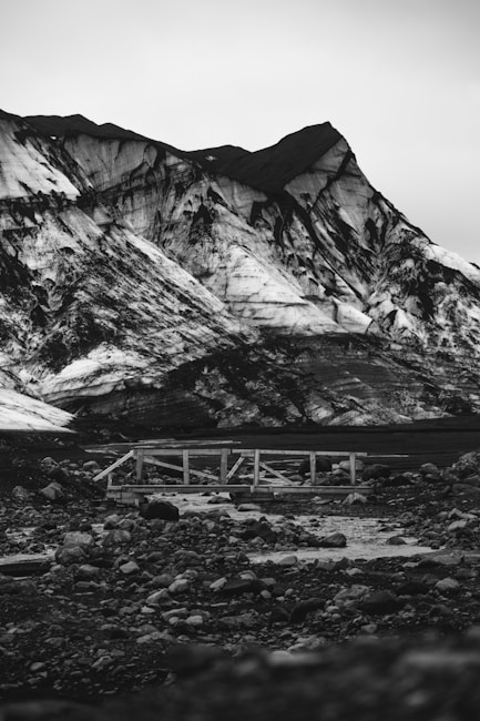 A dramatic black and white photograph of a rugged mountainous landscape with prominent geological features. The imposing mountain peaks are sharply defined against the sky, with intricate patterns of light and shadow playing across their faces. A small wooden bridge extends over a rocky stream in the foreground, adding a sense of scale and depth.