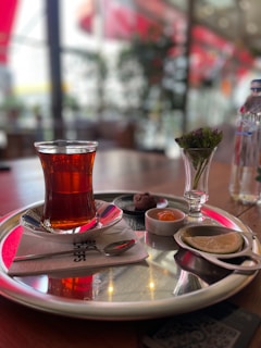 A glass of tea is served on a silver tray, accompanied by a small dish of chocolates, a slice of lemon, and a small vase with flowers. The setting suggests a cafe or restaurant environment, with blurred background elements and soft lighting enhancing the atmosphere.