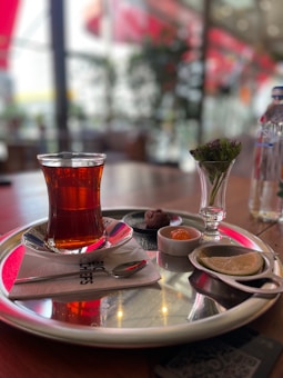 A glass of tea is served on a silver tray, accompanied by a small dish of chocolates, a slice of lemon, and a small vase with flowers. The setting suggests a cafe or restaurant environment, with blurred background elements and soft lighting enhancing the atmosphere.