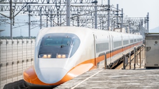 A high-speed train is positioned on a rail track beneath a structure lined with numerous overhead power lines. The sleek, aerodynamic front of the train features a white and orange color scheme with reflective windows. The train platform is visible, emphasizing the advanced rail infrastructure.