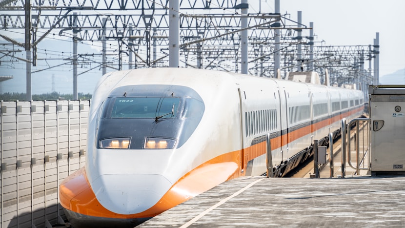 A high-speed train is positioned on a rail track beneath a structure lined with numerous overhead power lines. The sleek, aerodynamic front of the train features a white and orange color scheme with reflective windows. The train platform is visible, emphasizing the advanced rail infrastructure.