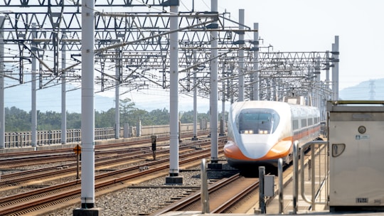 A sleek, modern high-speed train moves along multiple railway tracks under a complex web of metal overhead structures. The train features a streamlined design with a white and orange color scheme. Surrounding the tracks are tall metal poles and electrical wires. The background includes distant greenery and hills under a light, clear sky.