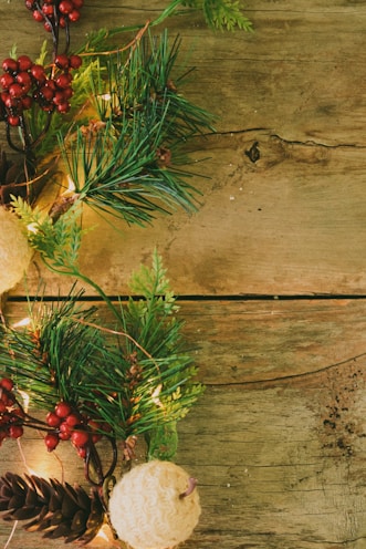 A rustic wooden surface decorated with festive elements, including pine branches, red berries, pinecones, and illuminated string lights intertwined with green foliage. The focus is primarily on the left side, with an emphasis on natural and cozy textures.