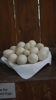 A collection of white eggs is piled in a bowl lined with a white paper towel, set against a wooden background. A label partially visible at the bottom suggests these might be owl eggs.