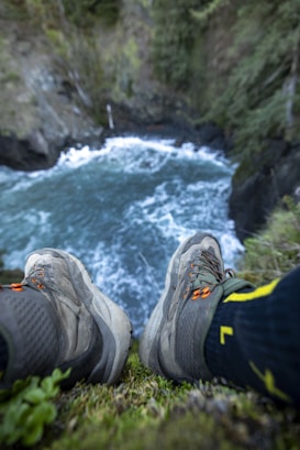 A pair of hiking boots is perched on the edge of a cliff above a flowing river. The scene is surrounded by lush greenery, with trees and rocks forming the backdrop. The water appears fast-moving and turbulent, creating a sense of depth and adventure.