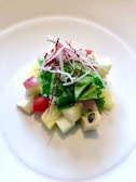 A colorful and vibrant salad arranged on a white plate. The salad consists of a mix of green leafy vegetables, small cubes of different fruits and vegetables such as apples and melons, as well as cherry tomatoes. On top, there are fine strips of radishes. The white plate and the natural lighting highlight the freshness of the ingredients.
