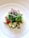 A colorful and vibrant salad arranged on a white plate. The salad consists of a mix of green leafy vegetables, small cubes of different fruits and vegetables such as apples and melons, as well as cherry tomatoes. On top, there are fine strips of radishes. The white plate and the natural lighting highlight the freshness of the ingredients.