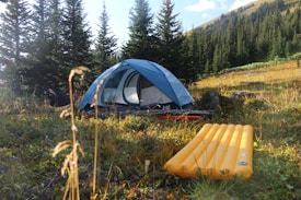A blue tent is set up in a grassy area surrounded by tall pine trees and rolling hills. In the foreground, there is a yellow inflatable sleeping pad. Sunlight filters through the trees, casting soft light on the camping setup, creating a peaceful outdoor atmosphere.